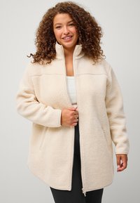 Giacca in pile beige chiaro con colletto alto, chiusura con zip e tasche laterali; texture morbida, design leggermente oversize.