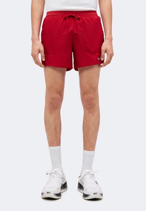Jambes masculines portant un short de running rouge Nike, des chaussettes blanches montantes et des chaussures de running blanches Nike, debout devant un fond uni.