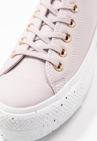 Zapatilla de piel de color rosa claro con puntera redonda, que presenta ojales dorados y cordones gris claro. Suela blanca texturizada con motas de colores.