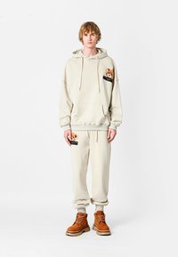 Conjunto de sudadera y pantalones beige fabricado con tejido suave, que presenta un parche de oso en el bolsillo y detalles en negro. Se lleva con botas marrones.