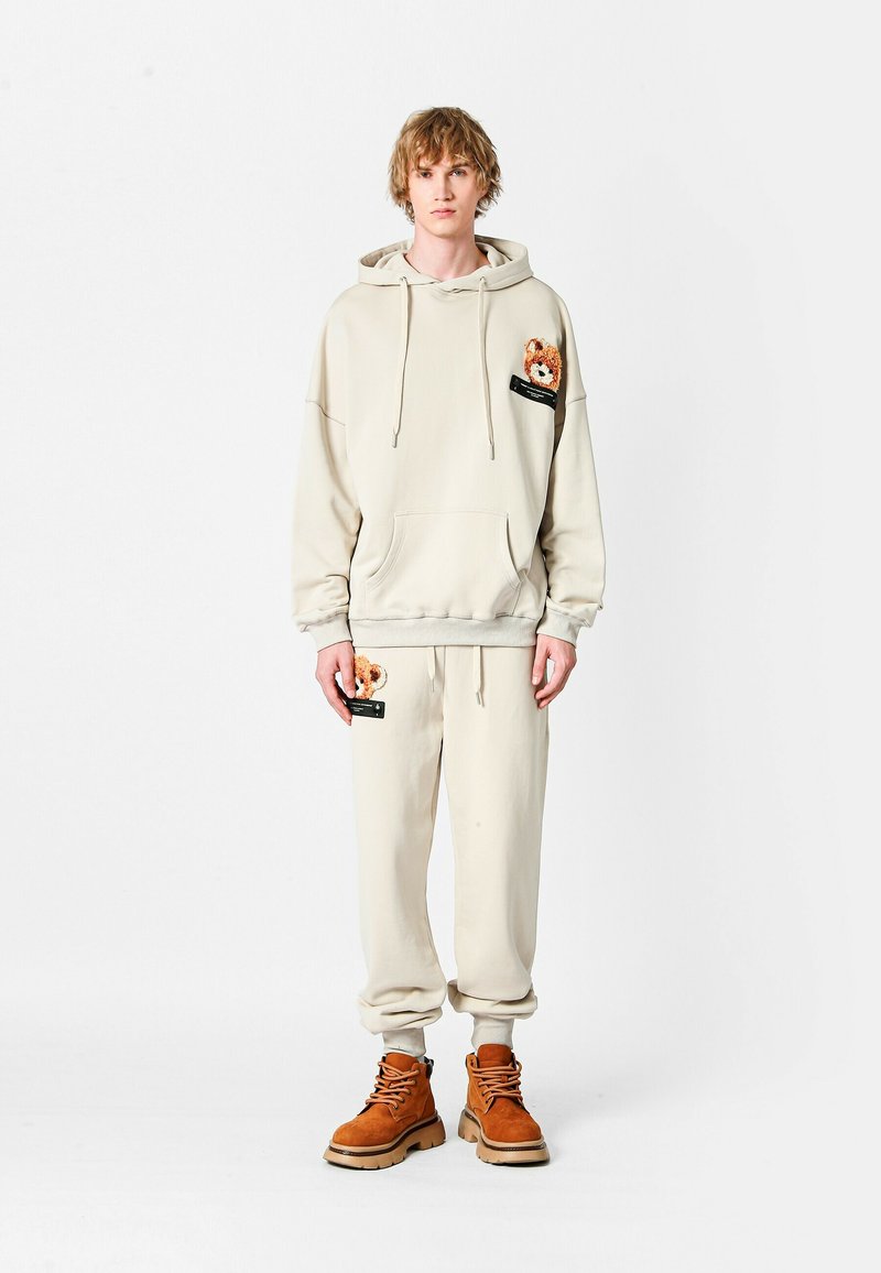 Conjunto de sudadera y pantalones beige fabricado con tejido suave, que presenta un parche de oso en el bolsillo y detalles en negro. Se lleva con botas marrones.