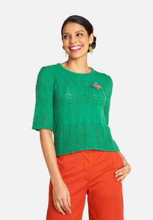 Lächelnde Frau, die ein grünes, kurzärmeliges Strickoberteil mit einer Blumennadel und leuchtend orangefarbenen Hosen trägt, mit einer Hand in der Tasche stehend.