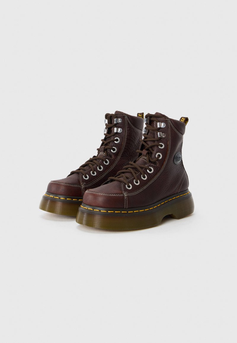 Bottes en cuir marron à lacets avec col rembourré, œillets métalliques, coutures jaunes et une semelle translucide épaisse. Présence du logo sur le côté.