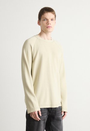 Light beige long-sleeve sweatshirt with a textured, ribbed fabric and raglan sleeves. Paired with dark jeans.