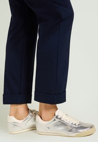 Zapatillas plateadas metálicas con detalles en crema, de perfil bajo y suela texturizada, combinadas con pantalones cortos azul marino remangados en los puños.