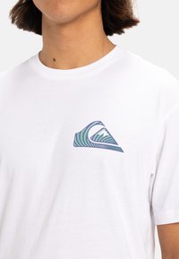 Camiseta blanca de algodón con un pequeño gráfico de ola colorida en el lado izquierdo del pecho, con rayas verdes y azules en forma triangular.