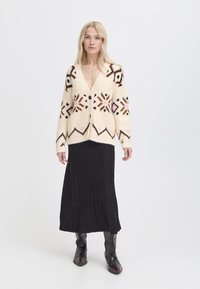 Cárdigan de lana gruesa con bordados geométricos en negro, rosa y marrón; combinado con una falda maxi negra de canalé y botas negras.