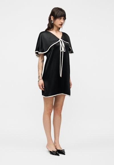 Μια γυναίκα στέκεται φορώντας μαύρο κοντό φόρεμα με λευκή επένδυση και λεπτομέρεια δέσιμου, μαύρα μυτερά παπούτσια χωρίς τακούνι και ασημένια σκουλαρίκια, μπροστά σε ένα απλό ανοιχτό φόντο.