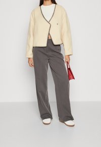 Kremowa kurtka z kontrastowym czarnym wykończeniem, szerokimi rękawami i dekoltem w serek; szare szerokie spodnie; czerwona torebka; białe buty.