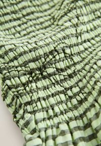 Tissu vert avec motif à carreaux noir, présentant une couture texturée élastique froncée créant une surface plissée.