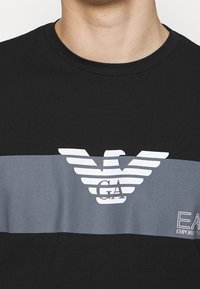 Černé bavlněné tričko s šedým horizontálním pruhem, na kterém se nachází bílé logo se stylizovanými křídly a textem "GA" a "EA" pod ním.