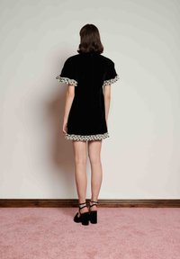 Γυναίκα που στέκεται αντιμετωπίζοντας έναν τοίχο, φορώντας ένα κοντό μαύρο φόρεμα με λευκή φόδρα που έχει πουά βολάν και μαύρα σανδάλια με ψηλά τακούνια, σε ροζ χαλί.