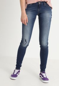 Jeans ajustados en denim azul oscuro, con un ligero desgaste en el muslo y la rodilla, combinados con zapatillas moradas y una blusa rosa claro.