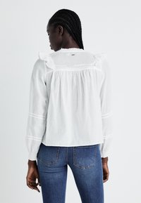 White long-sleeve blouse featuring ruffle accents, a loose fit, and delicate horizontal stripes. Paired with blue fitted jeans.