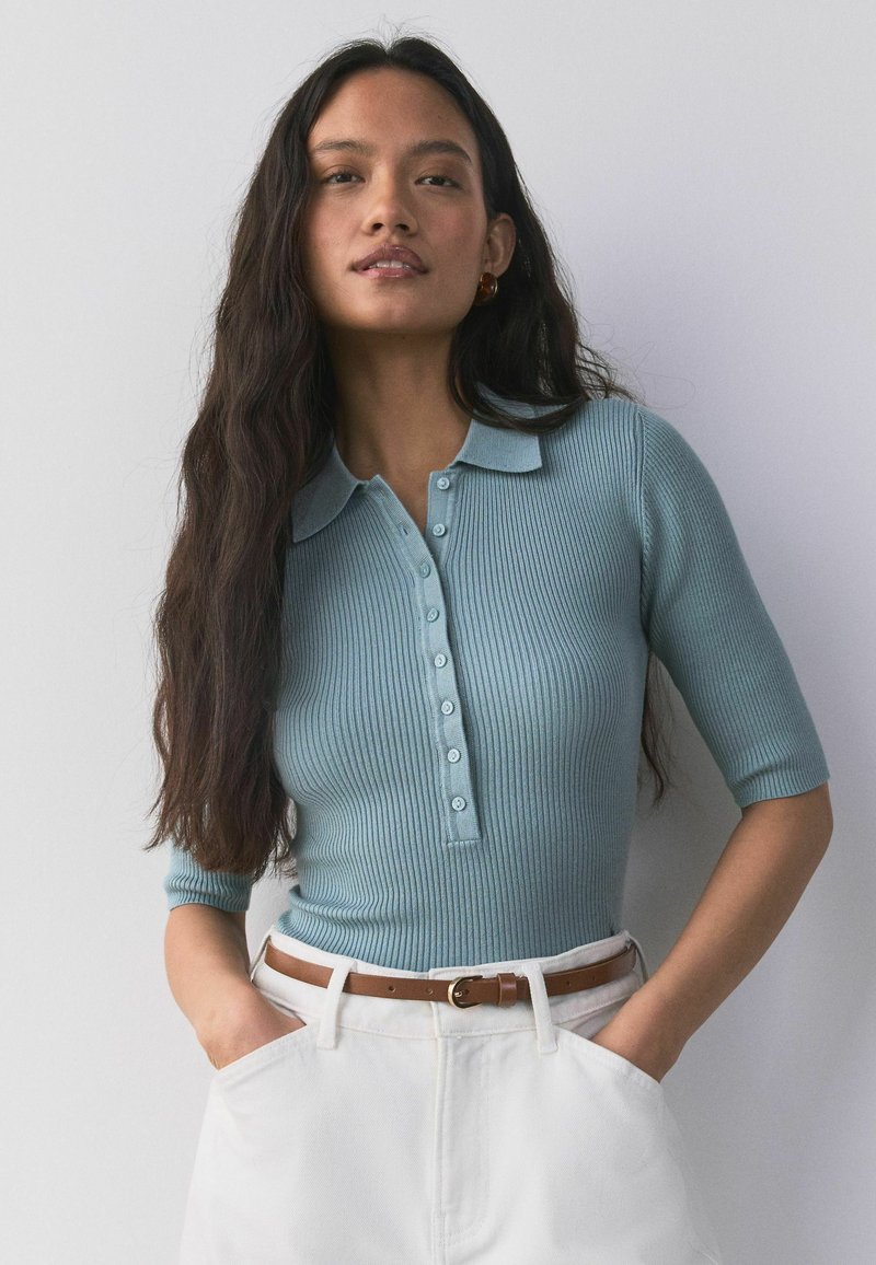 Woman with long dark hair wearing a light blue ribbed polo shirt and white pants with a brown belt, hands in pockets, against a plain background.