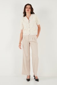 Ženska v belem teksturiranem kratkih rokavih žepni jopici s gumbi, bež širokih hlačah z visokim pasom in črnih koničastih petah z gležnjskimi trakovi.