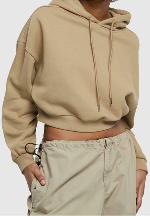 Osoba ubrana w beżową krótka bluzę z kapturem i sznurkami oraz jasnobeżowe luźne spodnie, z rękami w kieszeniach, na jednolitym tle.