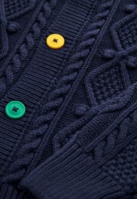 Sötétkék kötött szövet texturált kábelmintákkal, két gombbal, egy sárga és egy zöld, mindkettő „Little Bird” dombornyomott felirattal.