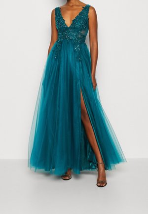 Kobieta w turkusowej sukni bez rękawów z kwiatowymi zdobieniami, tiulową spódnicą i rozcięciem sięgającym uda, w czarnych sandałach na paski.