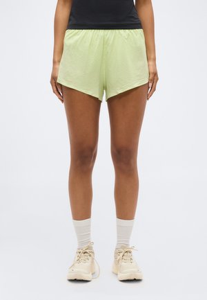 Γυναίκα που φοράει ανοιχτό πράσινο σορτς γυμναστικής, λευκές κάλτσες μέχρι τους αστραγάλους, μπεζ αθλητικά παπούτσια και μαύρο αμάνικο μπλουζάκι, στέκεται μπροστά σε ένα απλό φόντο.