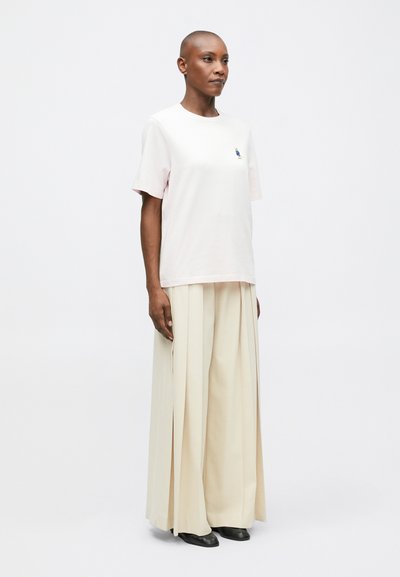 Osoba s krátkými vlasy, oblečená v bledě růžovém tričku a širokých krémových plisovaných kalhotách, stojící před jednobarevným bílým pozadím.