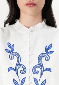 Camicia bianca con colletto arricciato, caratterizzata da motivi floreali ricamati blu e piccoli bottoni frontali. Tessuto di cotone liscio.