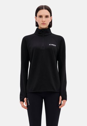 Frau trägt ein schwarzes langärmeliges Sportoberteil mit Reißverschluss und dem Logo „cep“ auf der Brust sowie schwarze Leggings mit dem Logo „60“ am Oberschenkel, steht frontal.