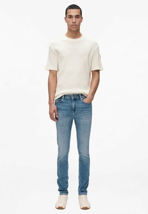 Un homme se tient devant un fond blanc, vêtu d’un tee-shirt blanc cassé, d’un jean bleu et de baskets blanches, une main dans la poche.
