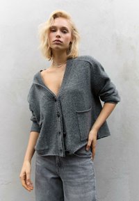 Siva kardigan iz mehkega blaga, z globokim V-izrezom, rebrastimi manšetami in sprednjim žepom, v kombinaciji s sivimi jeans hlačami.