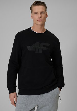 Mężczyzna w czarnej bluzie z logo "4F Sportswear" i jasnoszarych spodniach, stojący na jednolitym szarym tle.