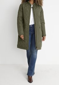 Abrigo largo acolchado en color verde oliva con cuello alzado, cremallera frontal y dos bolsillos laterales. Lleva puesto sobre una camisa blanca y unos jeans azules.