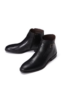 Bottes dcuir noir jusqu cheville avec une finition lisse, dotes dun panneau textur, dun fermoir  glissire latrale et dune bride dcorative avec boucle.