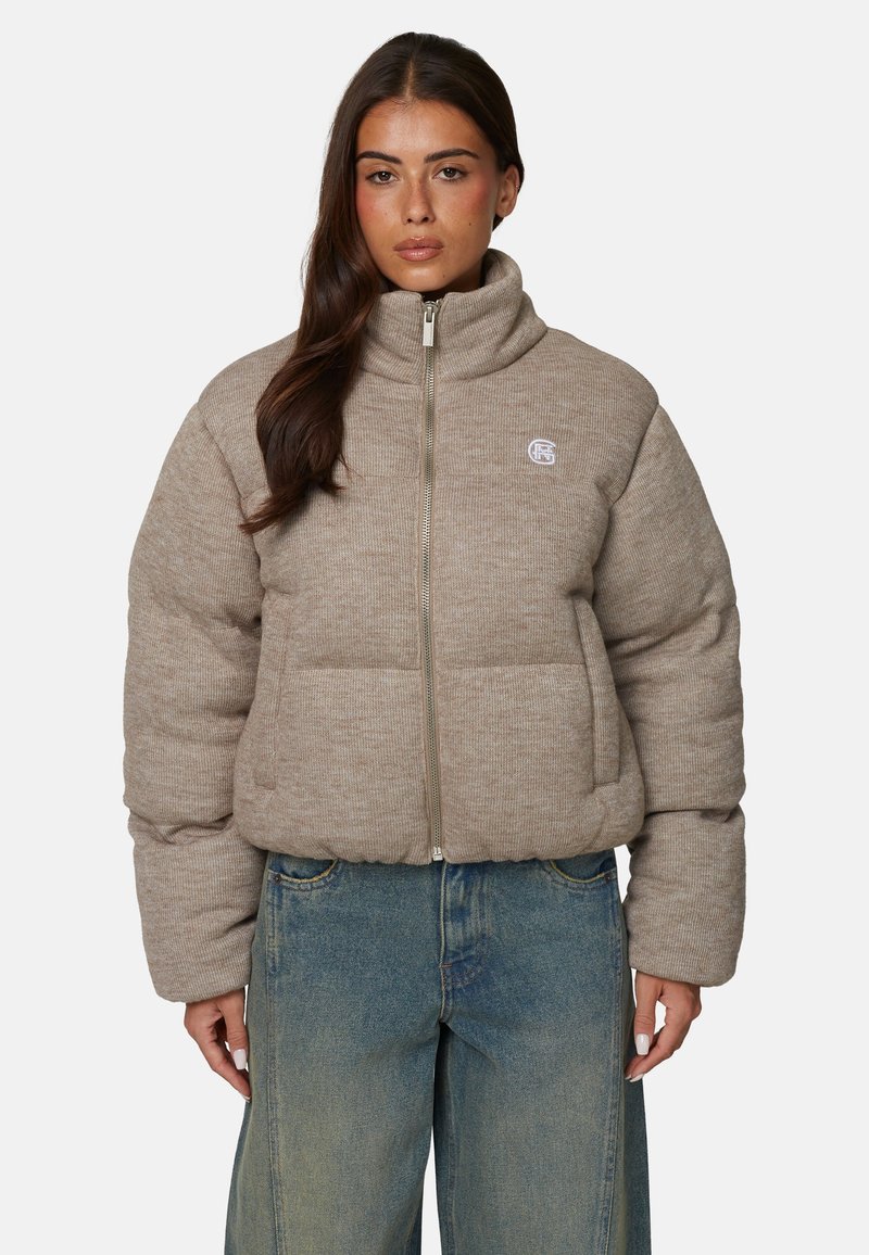 Beżowa puchowa kurtka z wysokim kołnierzem, zamek błyskawiczny i dużymi pikowanymi sekcjami. Ma małe logo na lewej piersi.