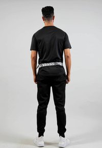 Uomo in piedi di spalle che indossa una maglietta nera, pantaloni neri, sneakers bianche e una cintura bianca con simboli neri.