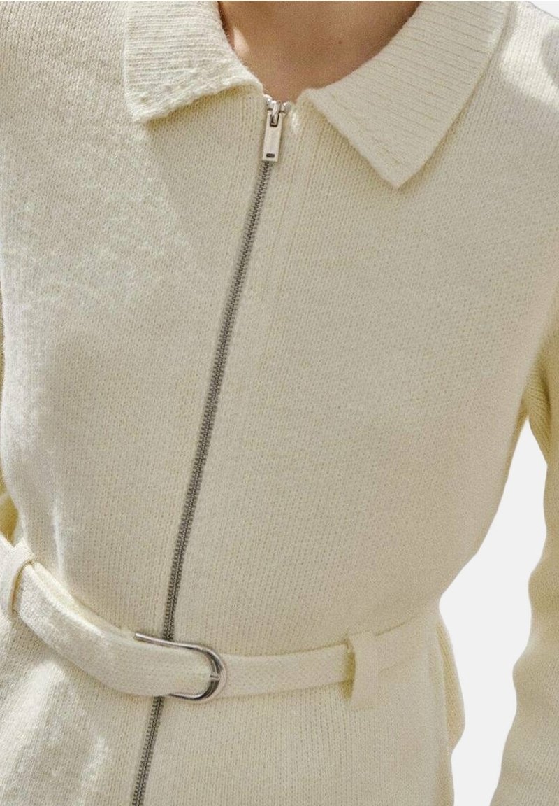 Cardigan en maille crème avec col, fermeture éclair métallique à l'avant et ceinture assortie avec boucle argentée à la taille.