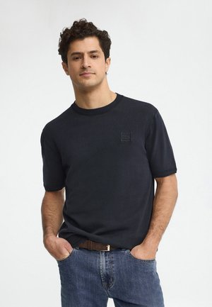 Uomo con capelli ricci che indossa un maglione nero a maniche corte e jeans blu, in piedi con le mani in tasca davanti a uno sfondo bianco.