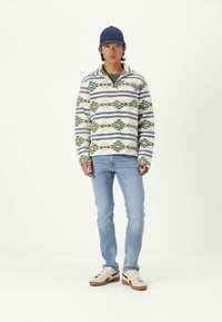 Giovane uomo che indossa un pullover di pile a fantasia, jeans azzurri, sneakers bianche e un cappellino da baseball blu navy, in piedi contro uno sfondo semplice.