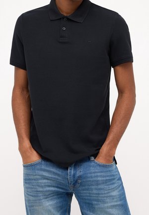 Mann trägt schwarzes Poloshirt mit kurzen Ärmeln und blaue Jeans, die Hände in den Taschen vor einfachem Hintergrund.