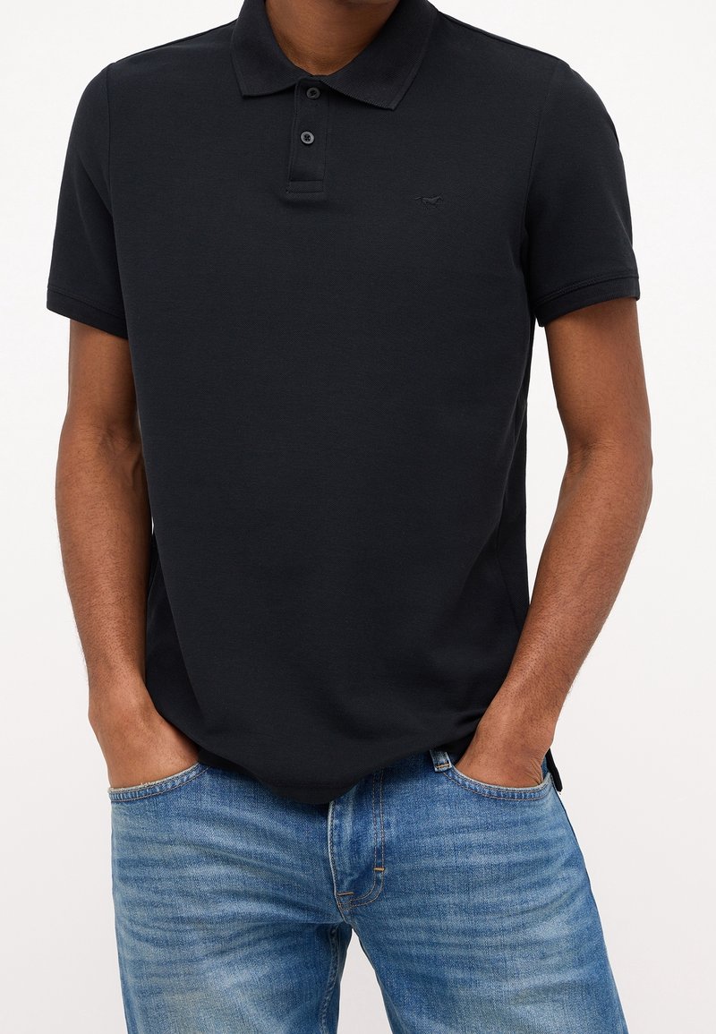 Homme portant un polo à manches courtes noir et un jean bleu, les mains dans les poches, sur un fond uni.