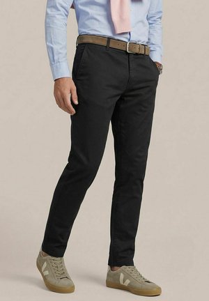 Man wearing black slim-fit pants, light blue shirt with pink sweater draped over shoulders, beige sneakers, and brown belt.