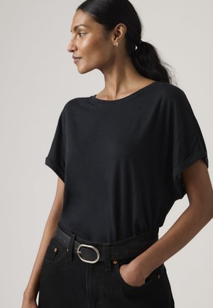 Γυναίκα με σκούρα μαλλιά σε χαμηλή αλογοουρά, φορώντας μια χαλαρή μαύρη μπλούζα που έχει μπει μέσα σε μαύρο παντελόνι με φαρδύ μαύρο ζωνάρι και ασημένιο αγκράφα.