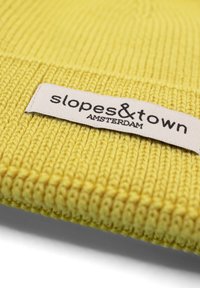 Gelbe gestrickte Mütze mit Rippenstruktur. Verfügt über ein rechteckiges weißes Etikett mit "slopes&town" und "AMSTERDAM" in schwarzer Schrift.