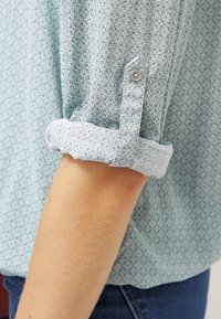 Chemise bleu clair avec un petit motif floral, manches retroussées avec un poignet blanc contrasté, en tissu léger, dotée d'une fermeture à boutons.