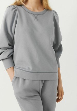 Kvinde iført lysegrå langærmet sweatshirt med pufskuldre og matchende sweatpants, hænderne i lommerne, mod en ensartet lys baggrund.