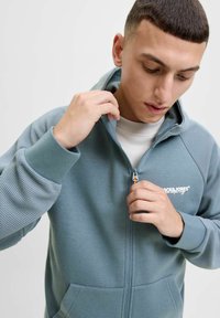 Sweatshirt zippé bleu clair avec un design texturé sur les manches, un logo sur la poitrine et des poches devant. Le tissu semble doux et lisse.