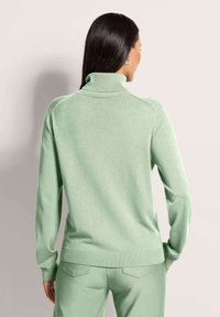 Frau mit langen dunklen Haaren, die einen hellgrünen Rollkragenpullover und passende Hosen trägt. Sie ist von hinten vor einem schlichten Hintergrund zu sehen.