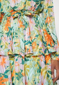 Blommig klänning med ett mönster i grönt, gult och orange. Har en samlad peplummidja och ett tygbälte. Lättviktsmaterial, genomskinligt.