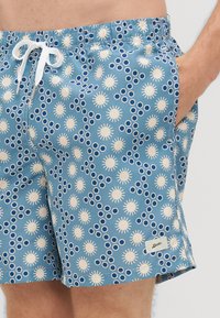 Zili peldēšanas šorti ar baltām saules un apļu rakstām, aukliņām jostas vietā, sānu kabatām un zīmolzīmi apakšmalā.