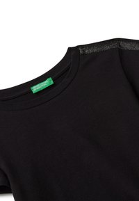 Černé tričko s kulatým výstřihem, s reflexním herringbone vzorem na rameni a zelenou etiketou United Colors of Benetton uvnitř límce.