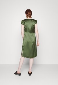 Robe en satin vert avec des manches courtes bouffantes, des fentes latérales et une longueur en dessous du genou. Portée avec des sandales à talons blocs noires.